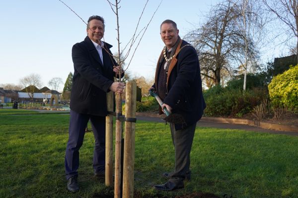 Two men in front of a tree, one is holding a spade full of dirt.