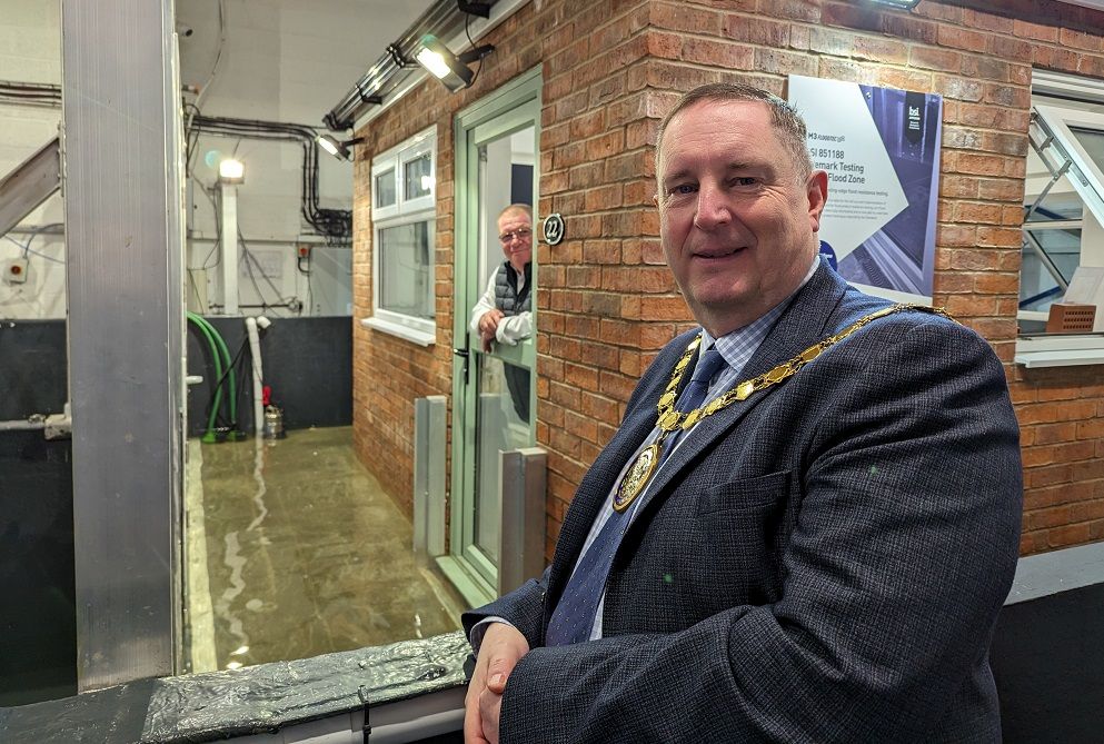 Cllr Robert Raphael leans on a gate with a house in the background where a man can be seen sticking his head out, while water pools around the house.