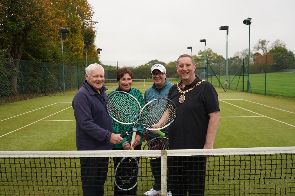 A group of people holding tennis rackets in front of a tennis net.