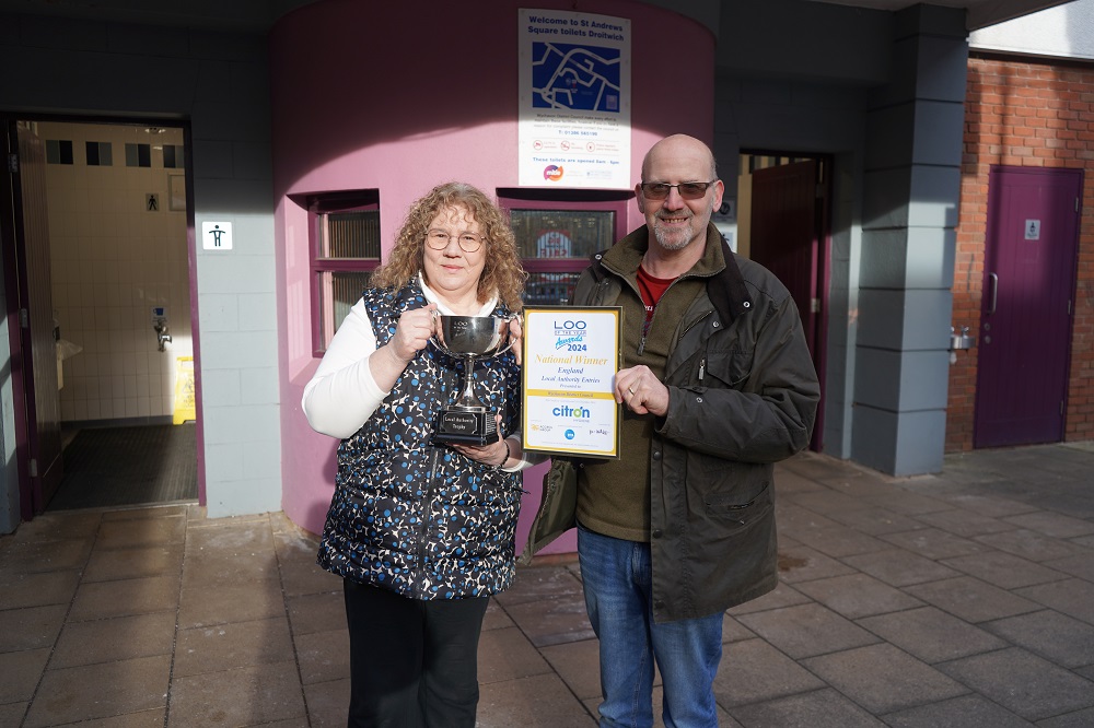 A woman holding a trophy and a man holding a certificate outside some toilets.