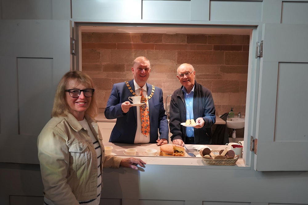 A woman stands in front of the hatch while Cllr Raphael and another man serve tea and cake