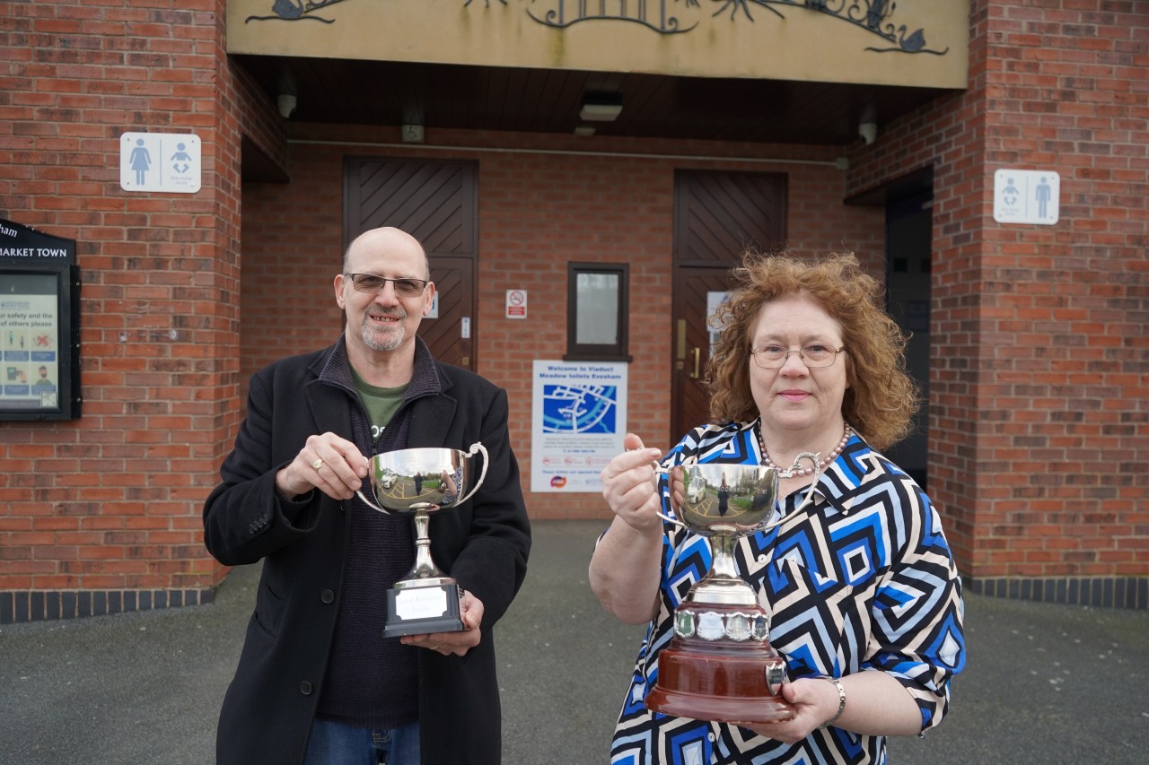 Phil Childs holding a trophy standing to the left, Cllr Emma Stokes holding a trophy and standing to the right outside a block of toilets.