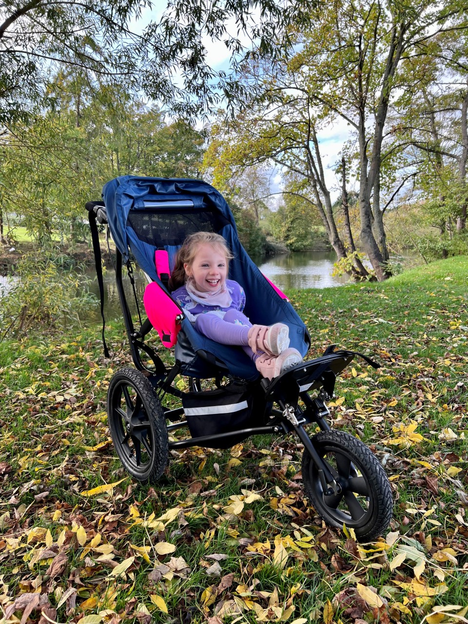 A beautiful young girl aged around six, looking very happy, sat in her pushchair surrounded by autumnal-looking background of trees and a river behind.